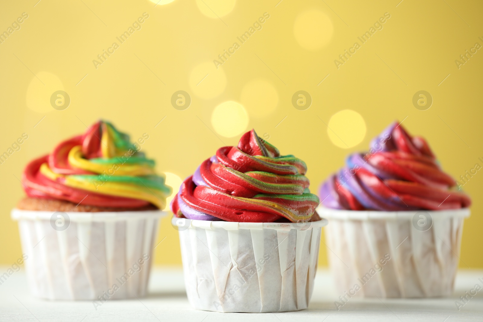 Photo of Delicious cupcakes with colorful cream on white table against golden background, closeup. Bokeh effect