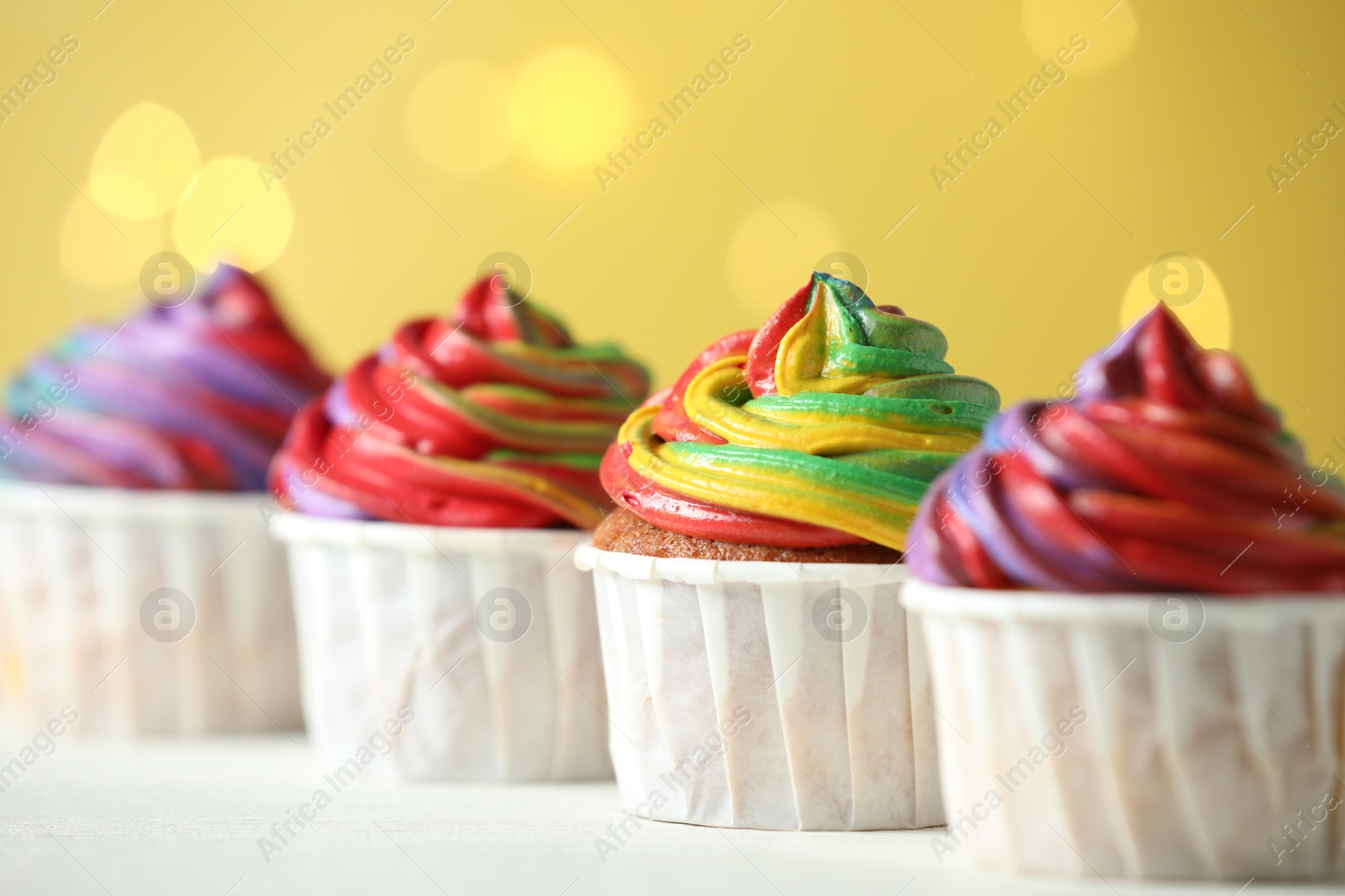 Photo of Delicious cupcakes with colorful cream on white table against golden background, closeup. Bokeh effect