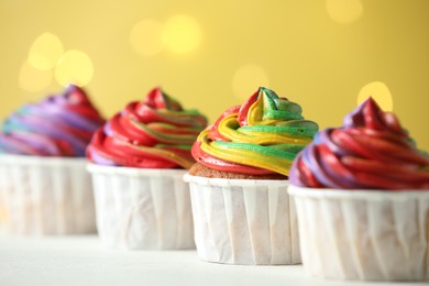 Photo of Delicious cupcakes with colorful cream on white table against golden background, closeup. Bokeh effect