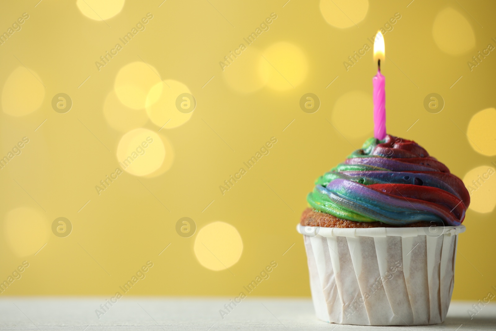 Photo of Delicious cupcake with colorful cream and burning candle on white table against golden background, closeup. Space for text