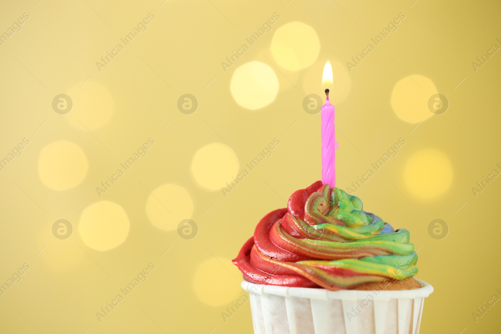 Photo of Delicious cupcake with colorful cream and burning candle against golden background, closeup. Space for text