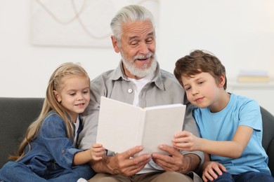 Photo of Grandpa and his grandkids reading book together on sofa at home