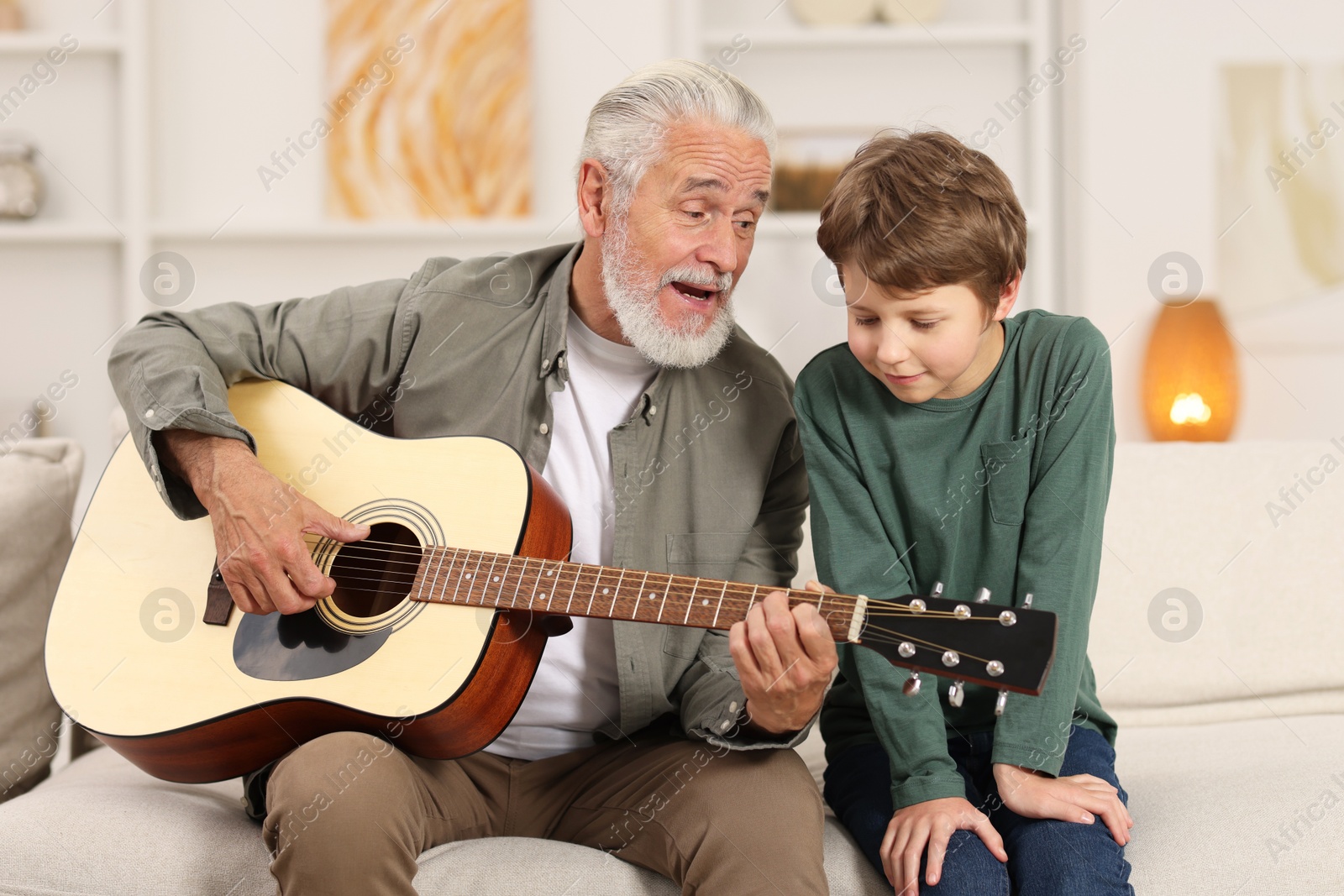 Photo of Grandpa teaching his grandson to play guitar on sofa at home