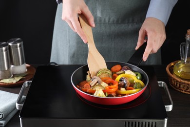 Photo of Woman frying vegetables and mushrooms in pan on stove at wooden table, closeup