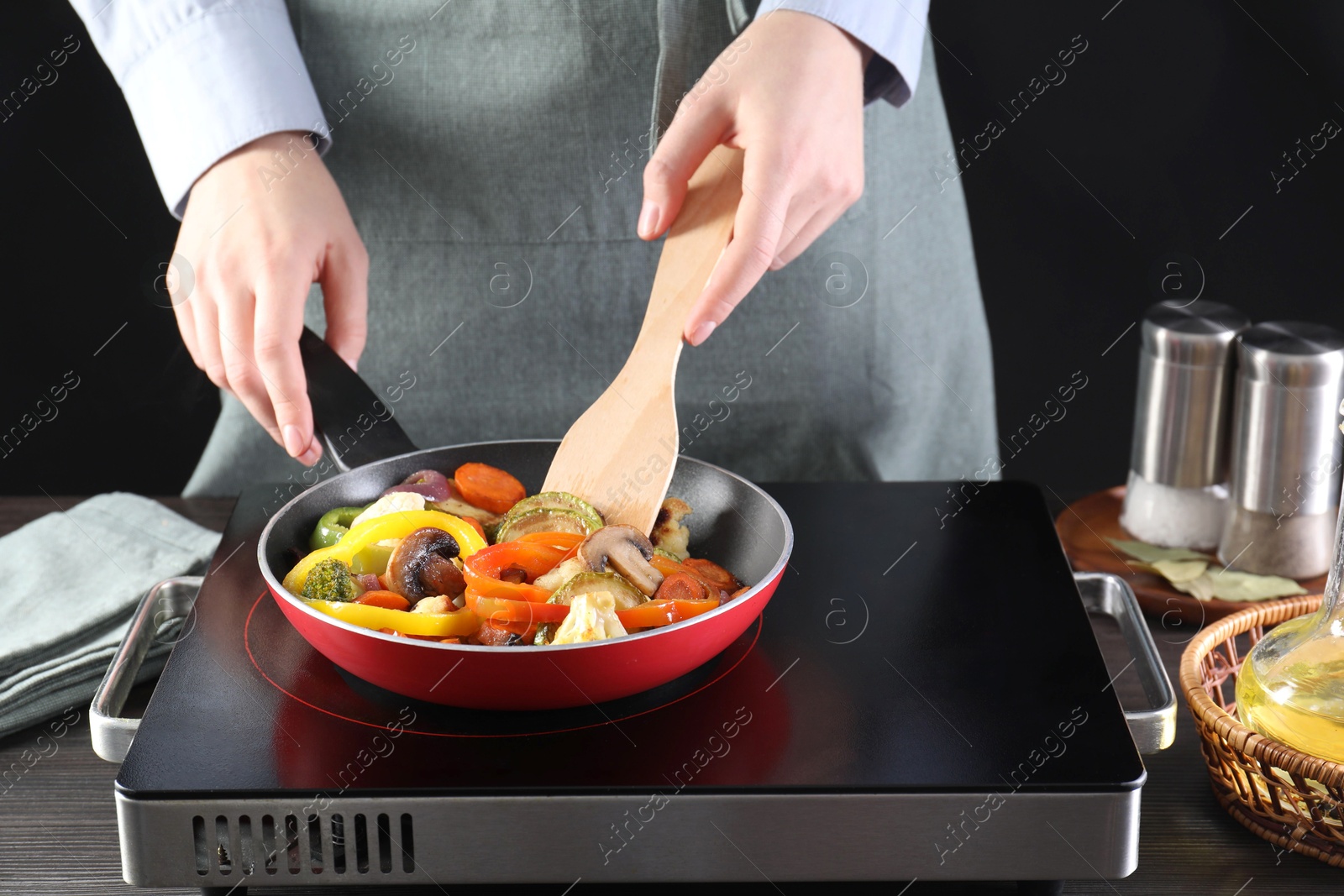 Photo of Woman frying vegetables and mushrooms in pan on stove at wooden table, closeup