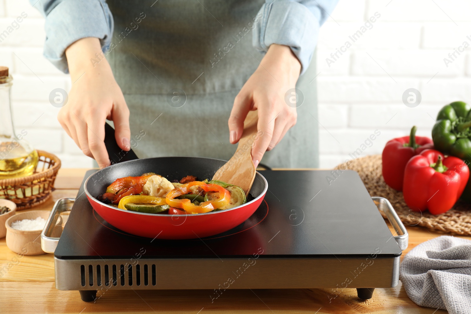 Photo of Woman frying vegetables in pan on stove at wooden table indoors, closeup