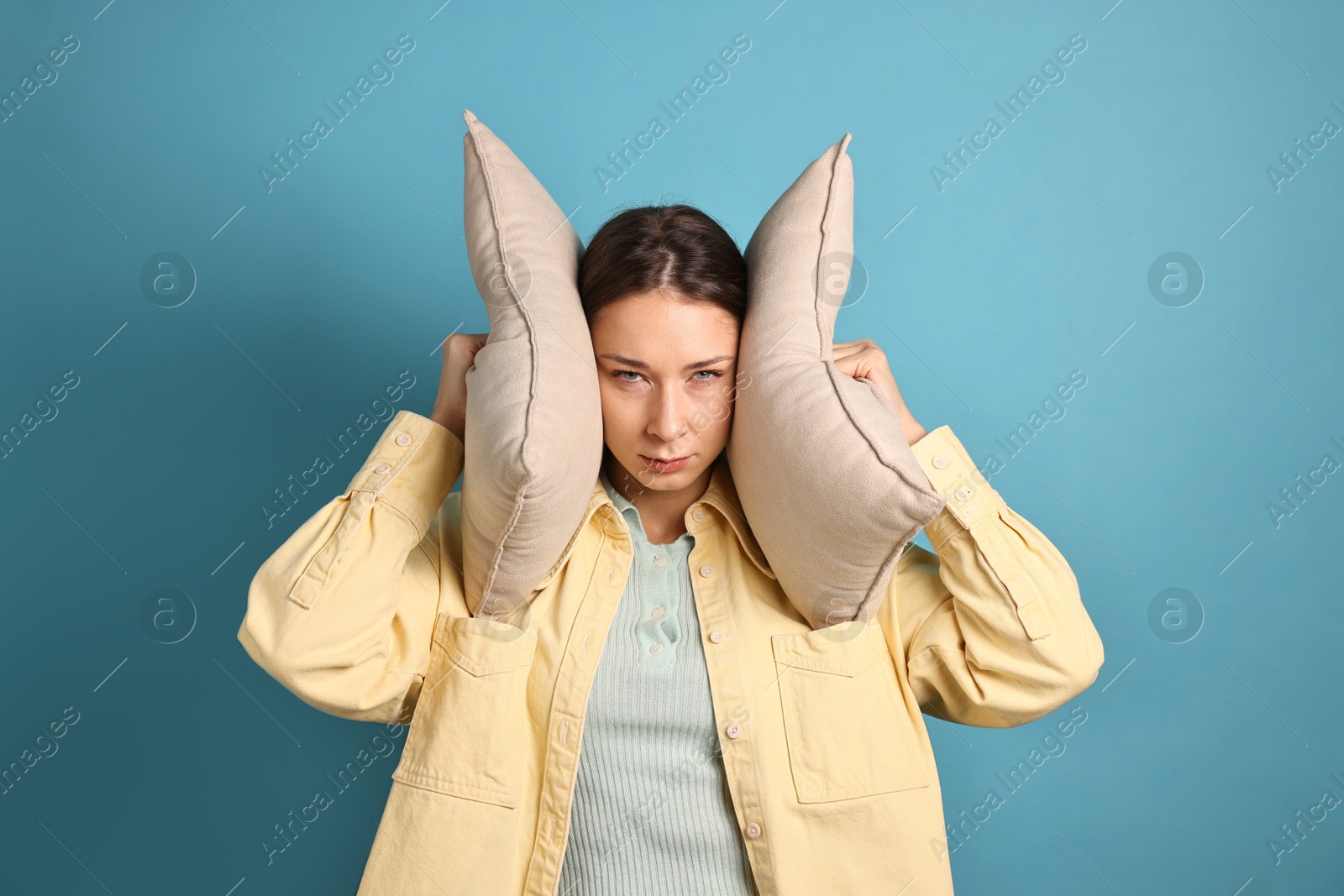 Photo of Annoyed woman covering her ears with pillows due to loud sound on light blue background