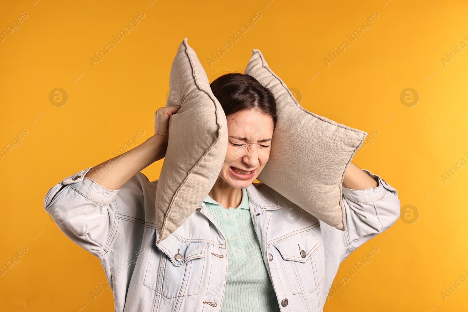 Photo of Annoyed woman covering her ears with pillows due to loud sound on orange background