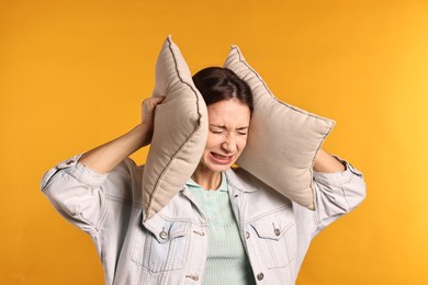 Annoyed woman covering her ears with pillows due to loud sound on orange background