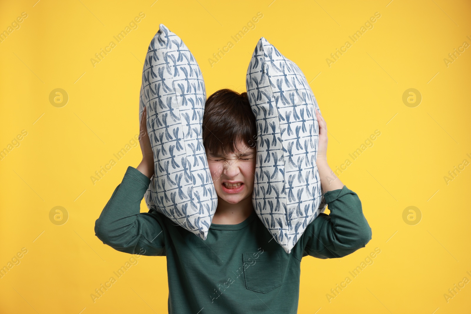 Photo of Annoyed boy covering his ears with pillows due to loud sound on yellow background