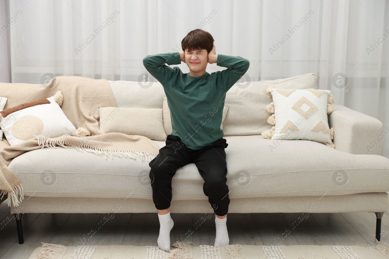 Photo of Annoyed boy covering his ears due to loud sound on sofa at home