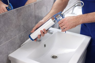 Photo of Worker with caulking gun sealing washbasin in bathroom, closeup