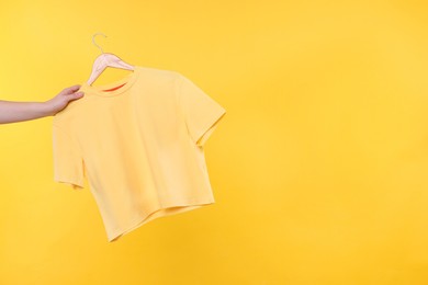 Photo of Woman holding hanger with t-shirt on yellow background, closeup. Space for text