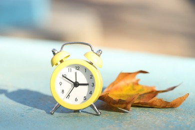 Photo of Autumn time. Alarm clock and golden leaf on light surface outdoors, closeup