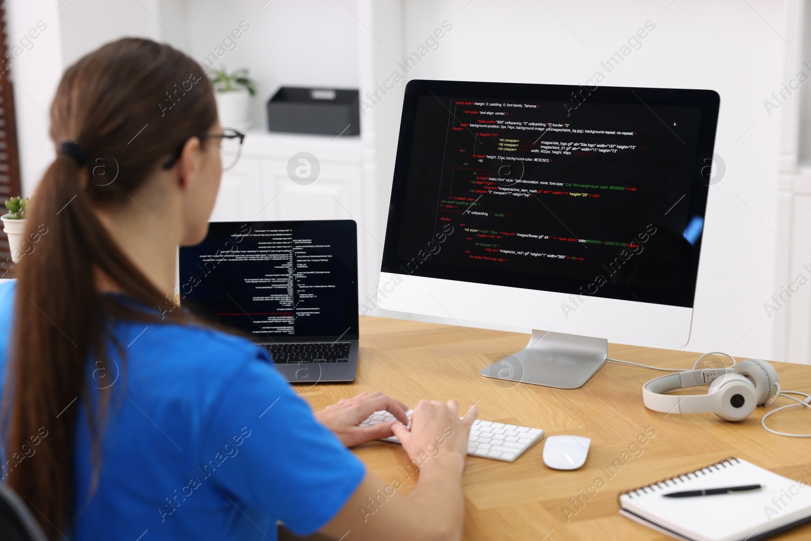 Photo of Programmer working on laptop and computer at wooden desk indoors, back view