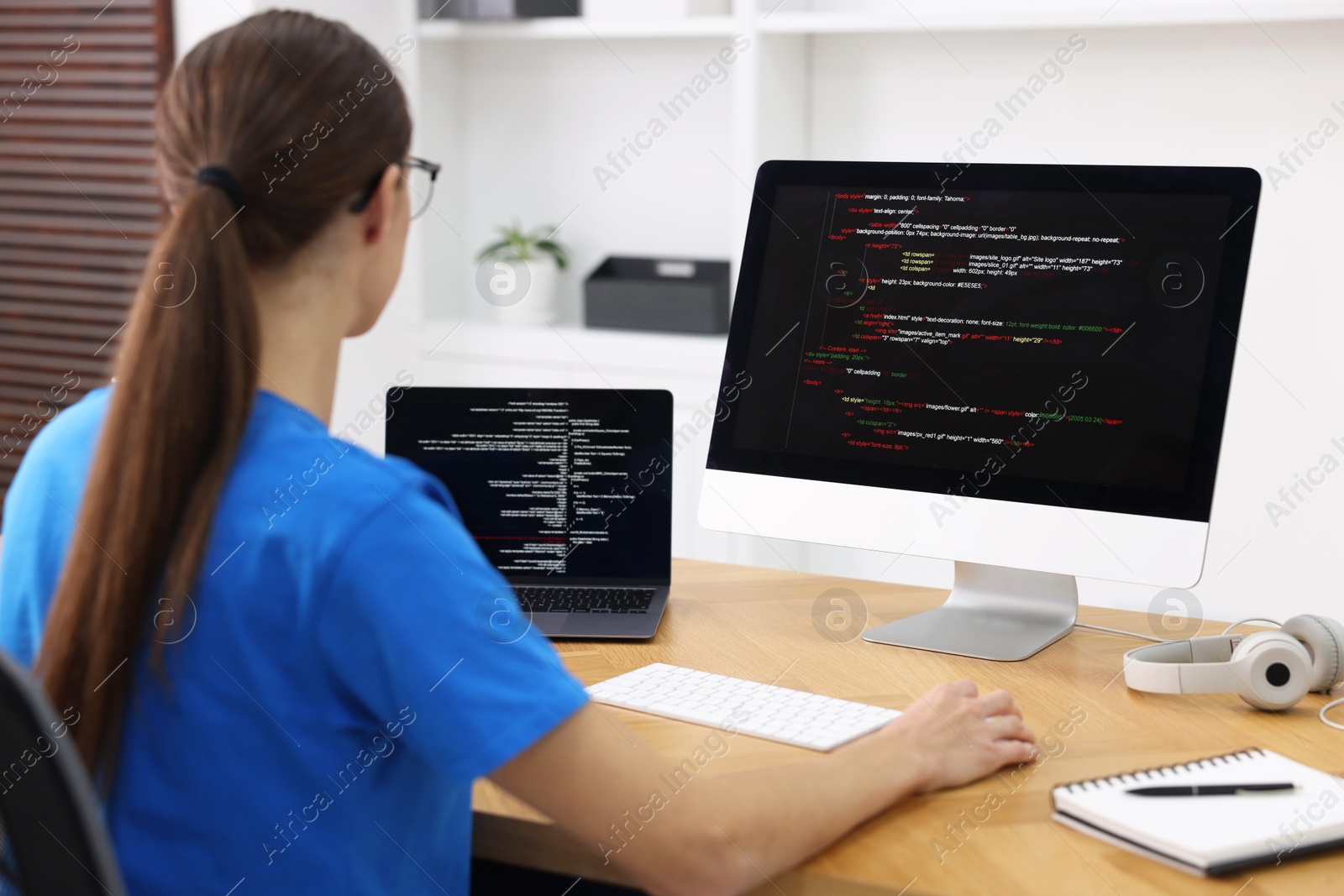 Photo of Programmer working on laptop and computer at wooden desk indoors, back view