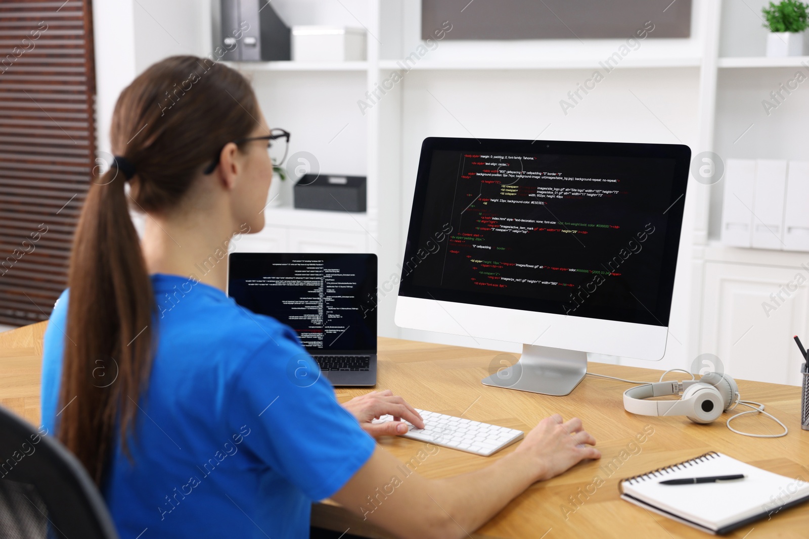 Photo of Programmer working on laptop and computer at wooden desk indoors, back view