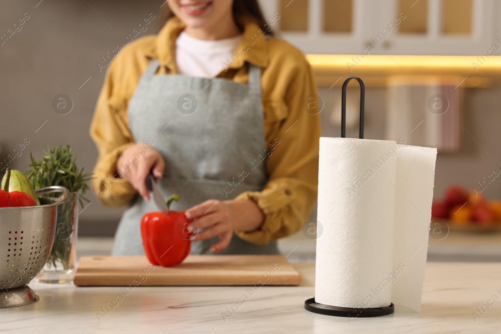 Photo of Woman cutting bell pepper at white marble table in kitchen, focus on roll of paper towels