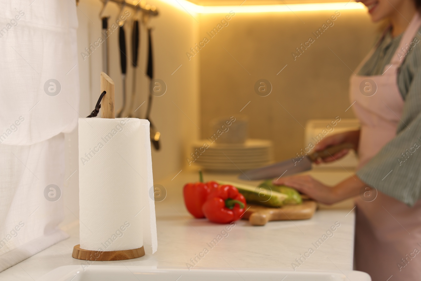 Photo of Woman cutting vegetables at white countertop in kitchen, focus on roll of paper towels