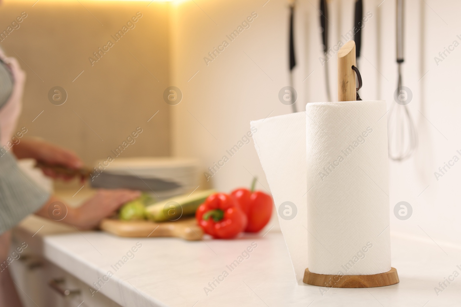 Photo of Woman cutting vegetables at white countertop in kitchen, focus on roll of paper towels