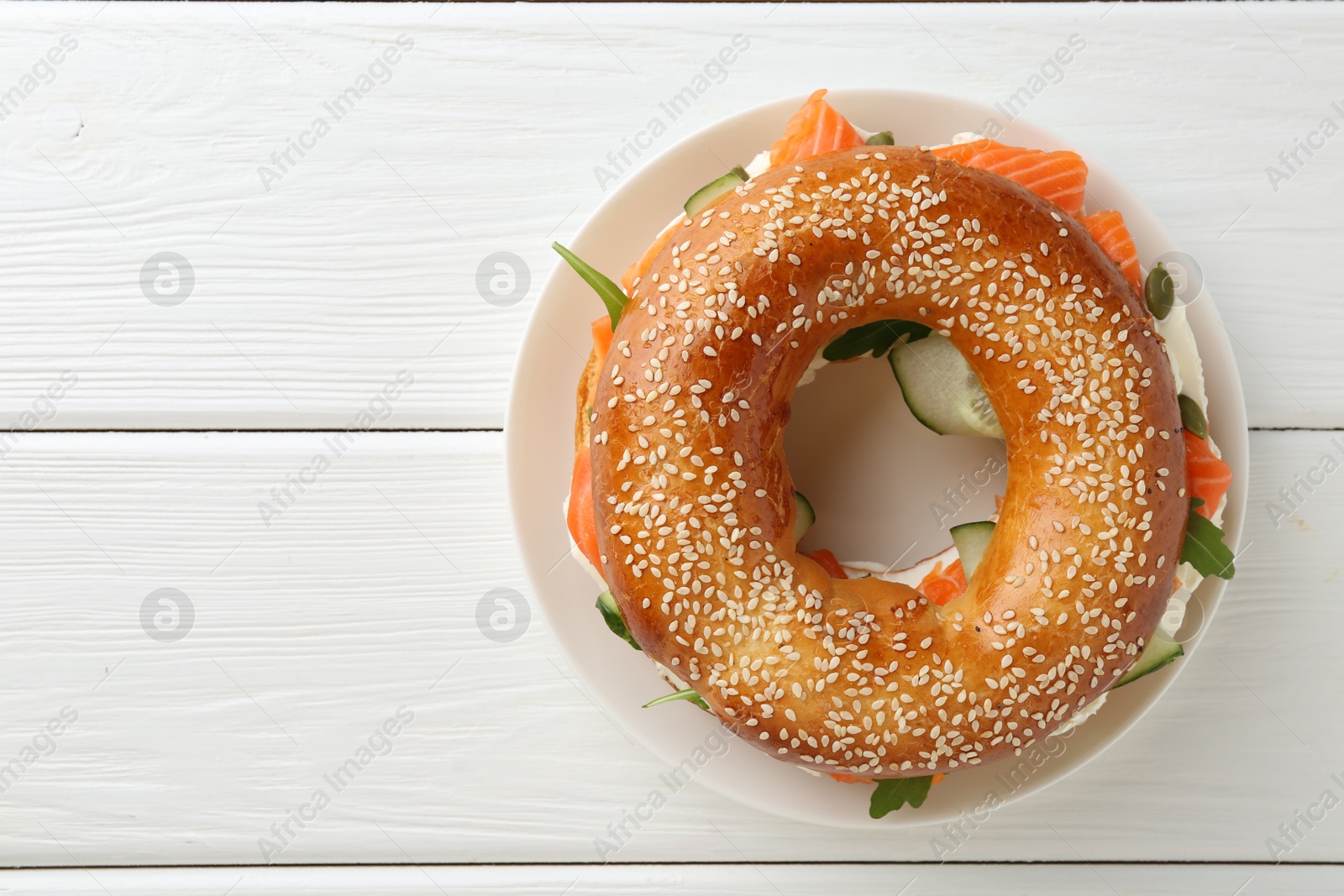 Photo of Delicious bagel with salmon, cream cheese and cucumber on white wooden table, top view. Space for text