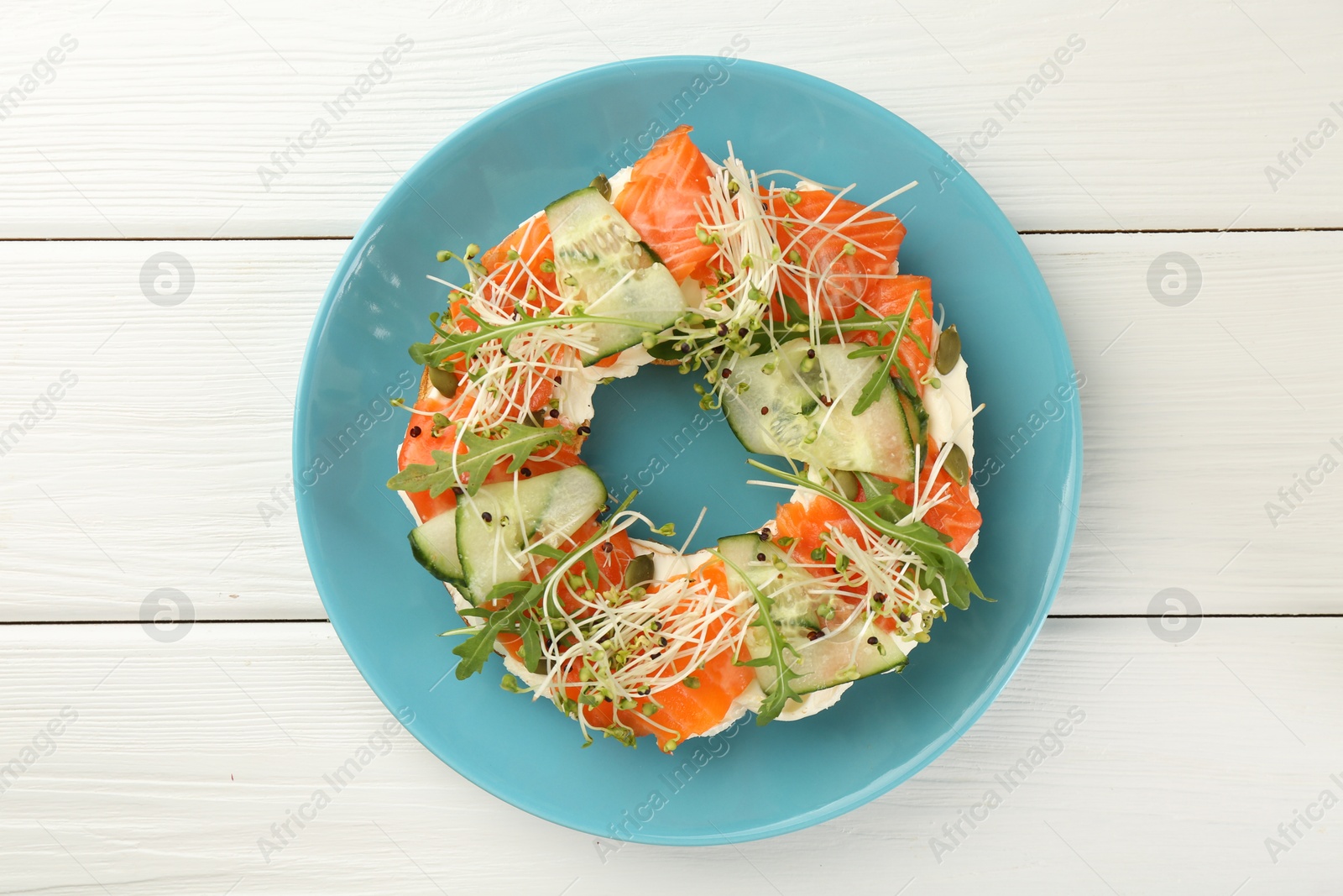 Photo of Half of delicious bagel with salmon, microgreens, arugula and cucumber on white wooden table, top view