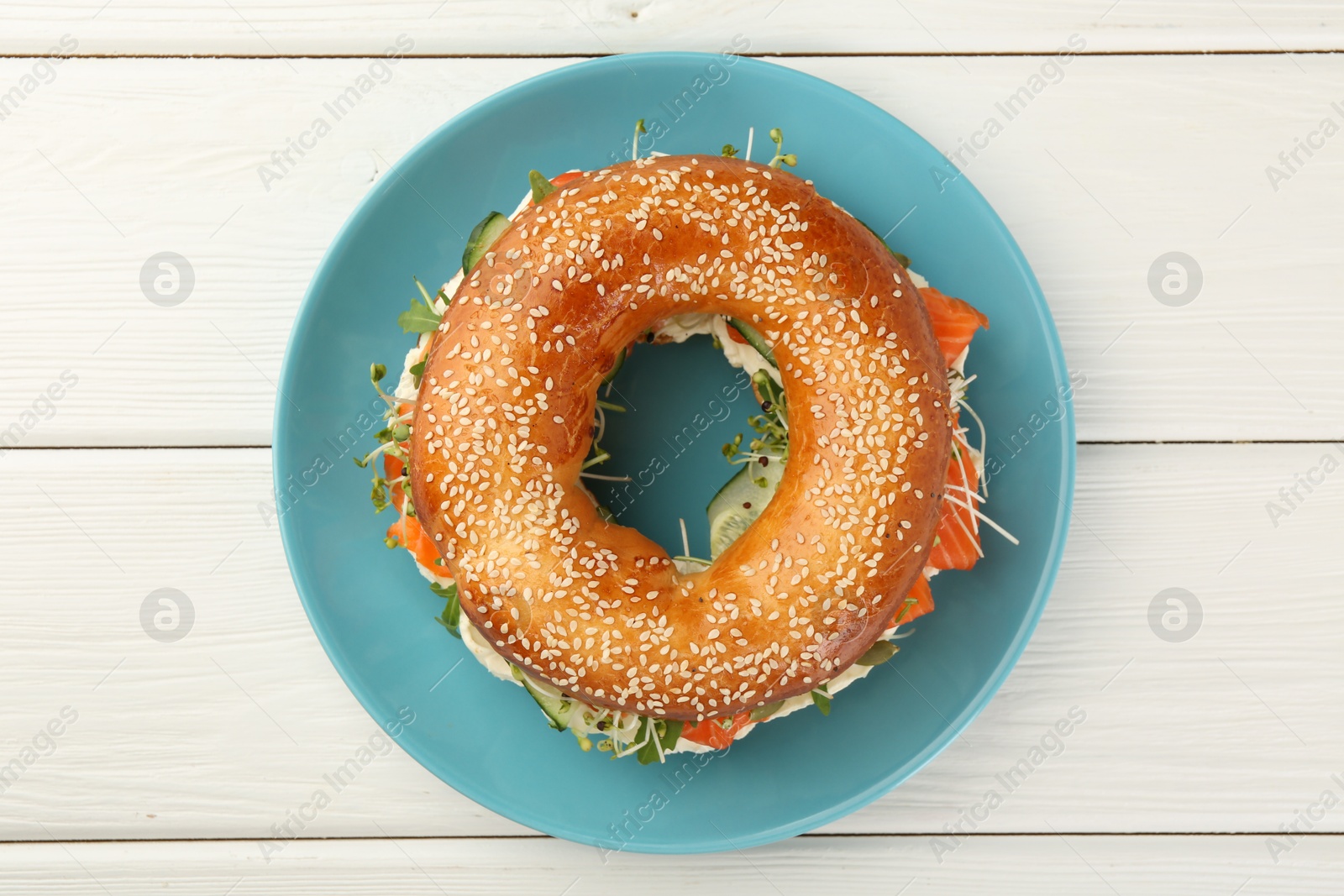 Photo of Delicious bagel with salmon, microgreens and cucumber on white wooden table, top view