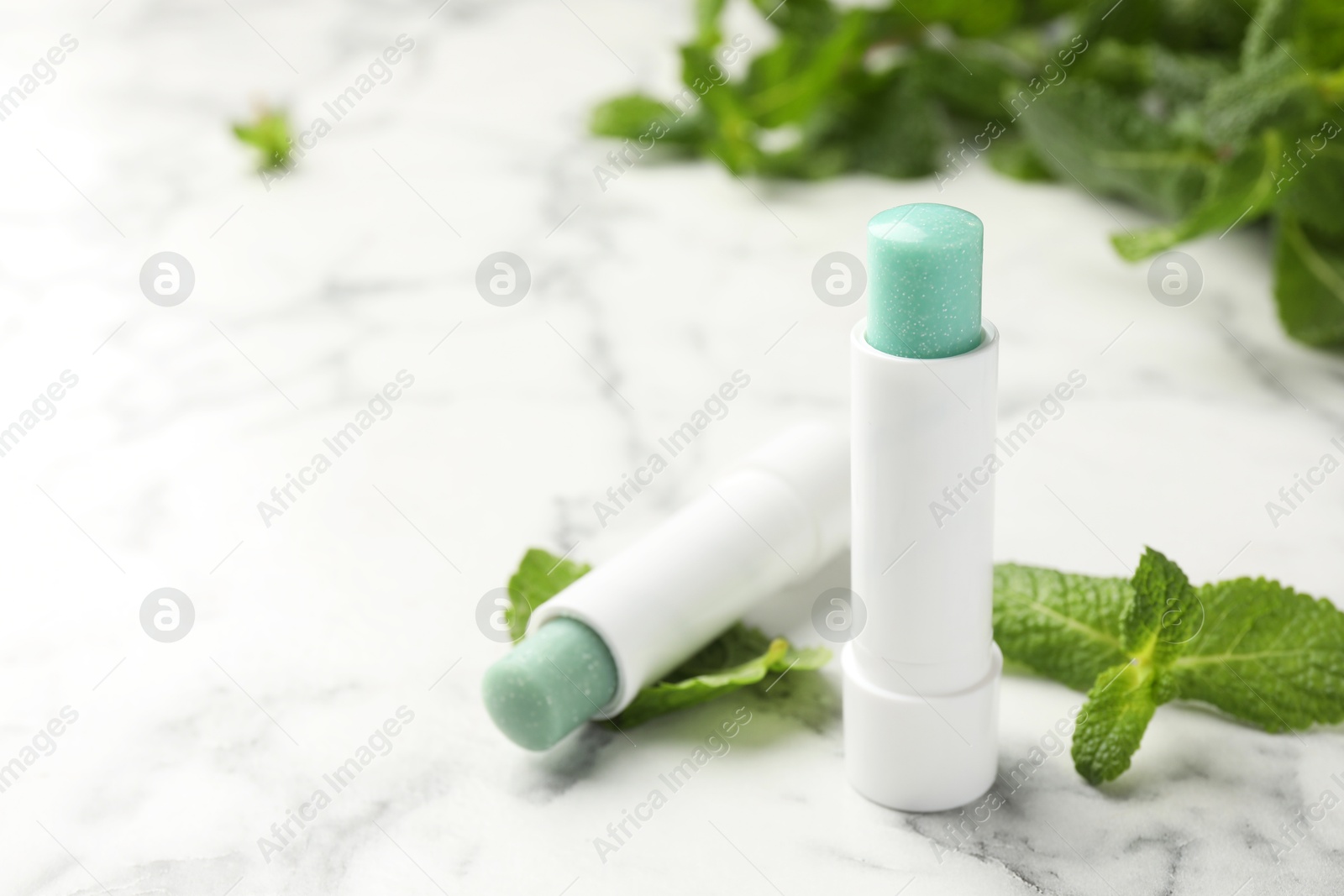 Photo of Natural lip balms and mint leaves on white marble background, closeup. Space for text