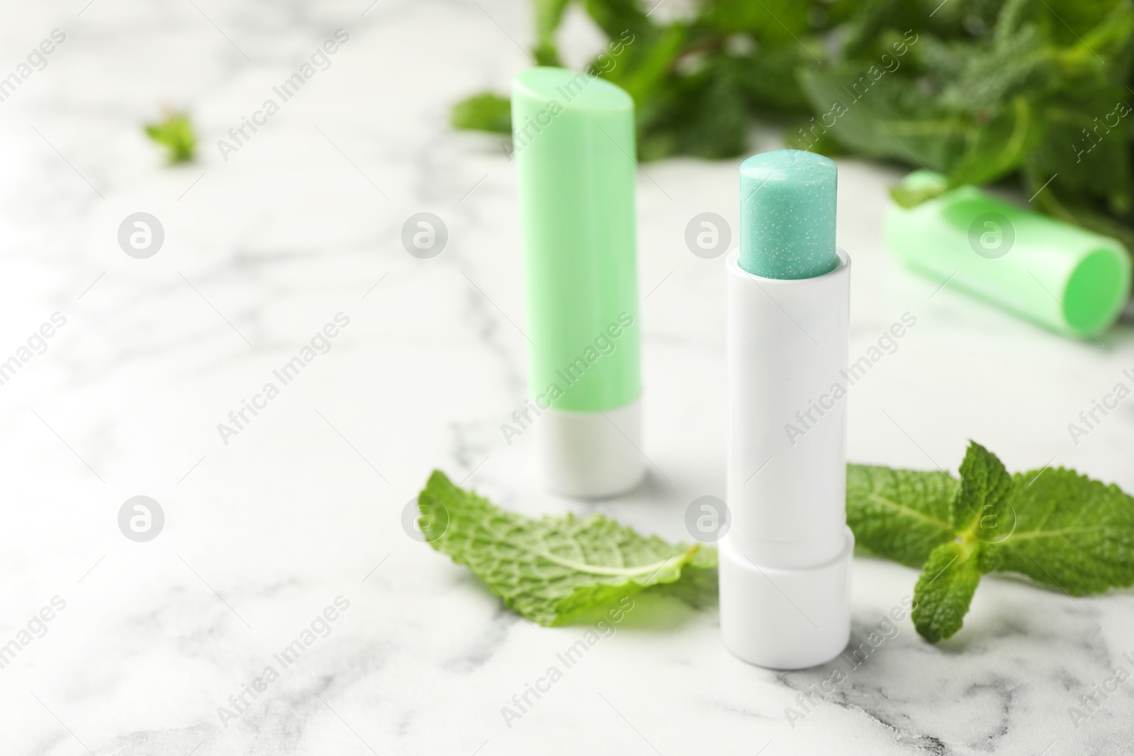 Photo of Natural lip balms and mint leaves on white marble background, closeup. Space for text