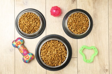 Photo of Dry pet food in feeding bowls and toys on wooden floor, flat lay