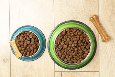 Photo of Dry pet food in feeding bowls and chew bones on wooden floor, flat lay