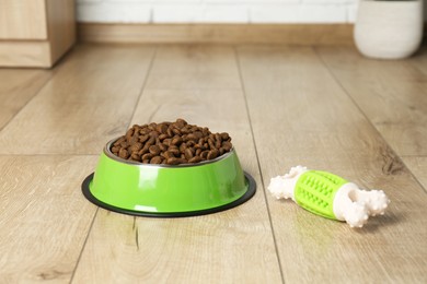 Photo of Dry pet food in feeding bowl and chew bone on floor indoors