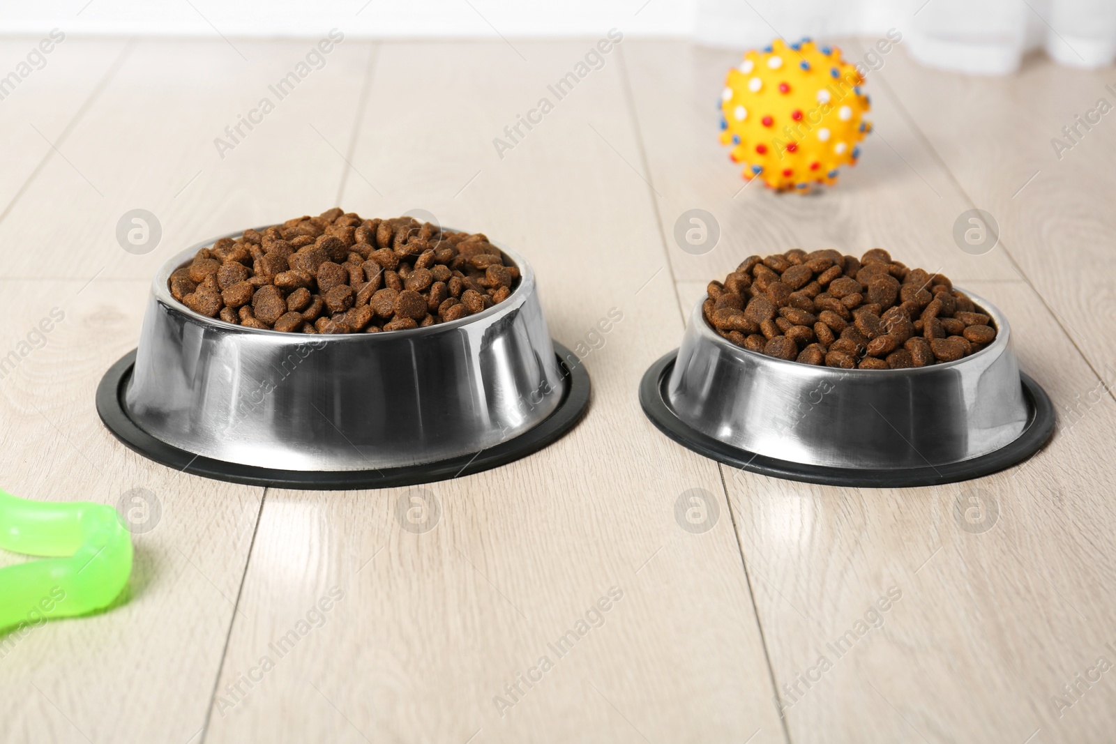 Photo of Dry pet food in feeding bowls and toys on floor indoors