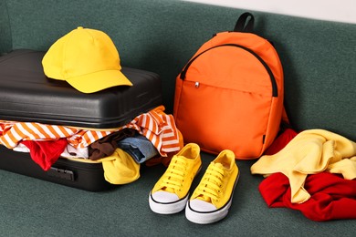Photo of Open suitcase full of different messy clothes, backpack and sneakers on sofa