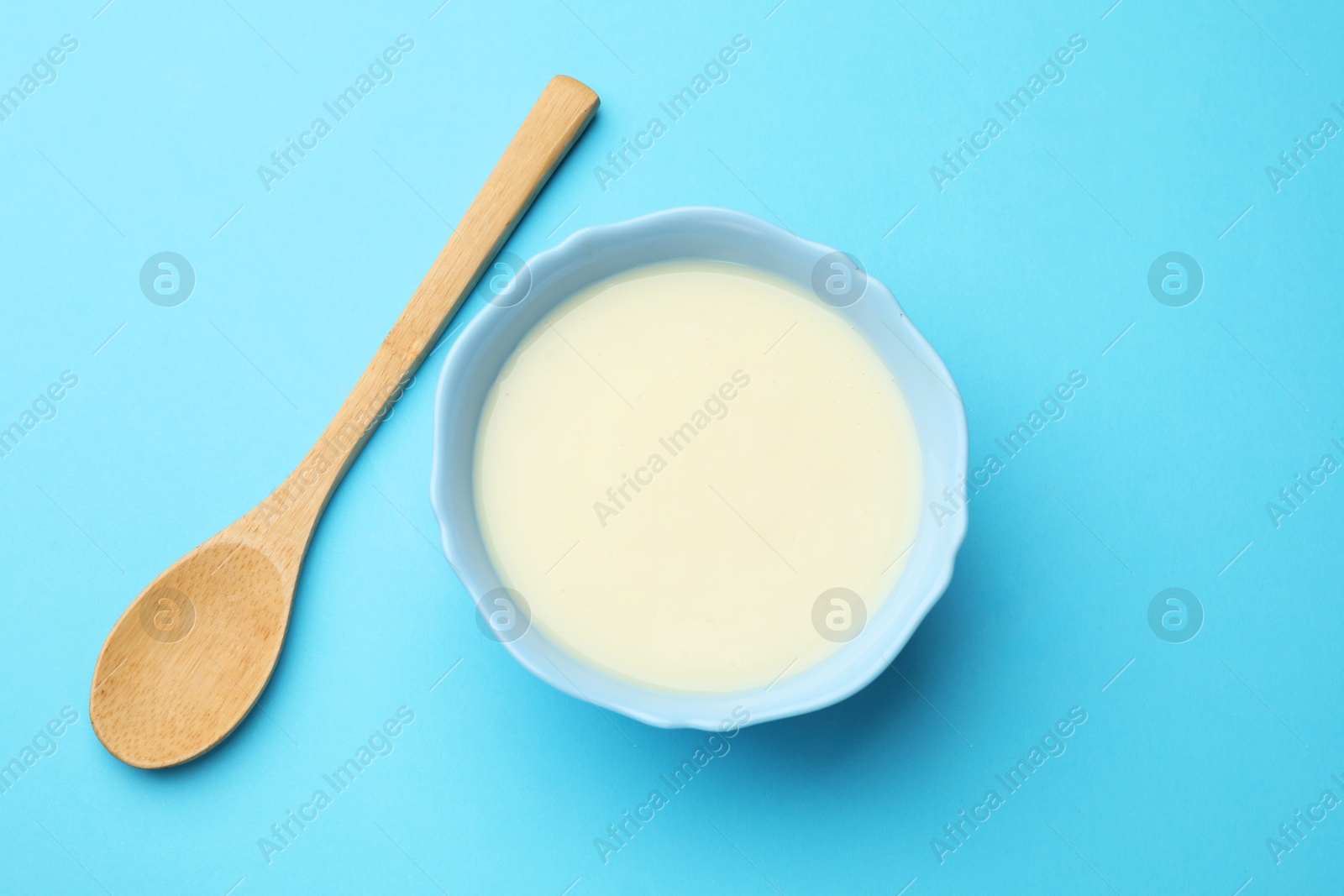 Photo of Tasty condensed milk in bowl and spoon on light blue background, top view