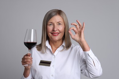 Senior woman with glass of wine showing okay gesture on grey background