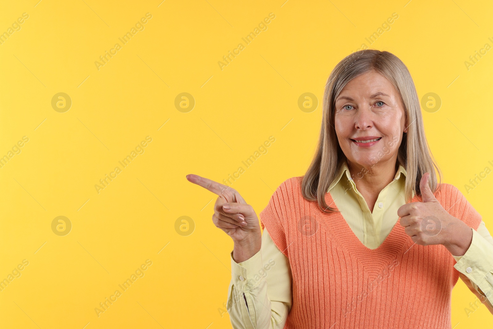 Photo of Senior woman showing thumbs up while pointing at something on yellow background. Space for text