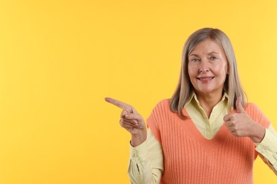 Photo of Senior woman showing thumbs up while pointing at something on yellow background. Space for text