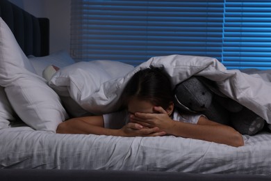 Photo of Fearful girl with toy bunny under duvet in bed at night