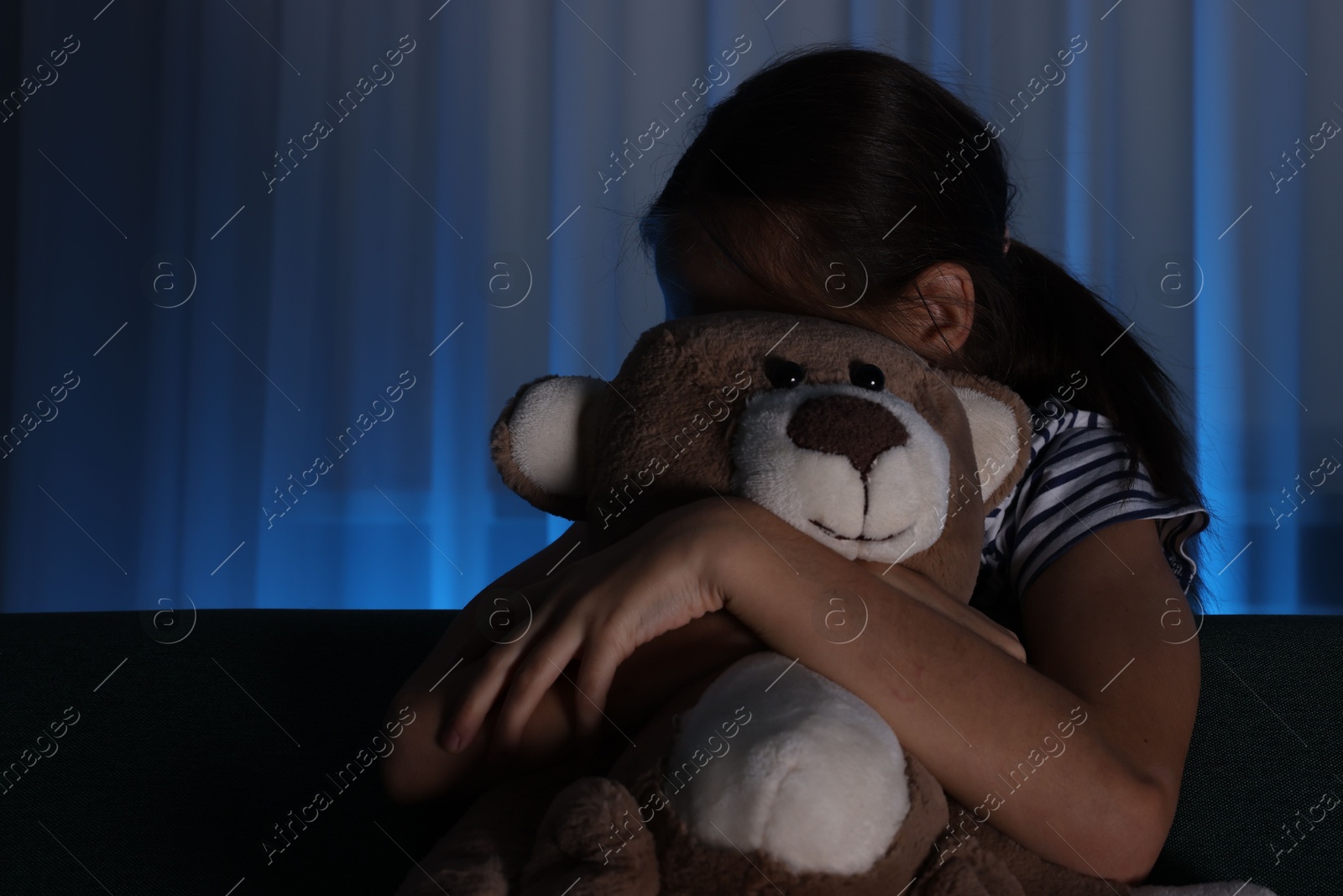 Photo of Scared girl with teddy bear hiding behind sofa at night