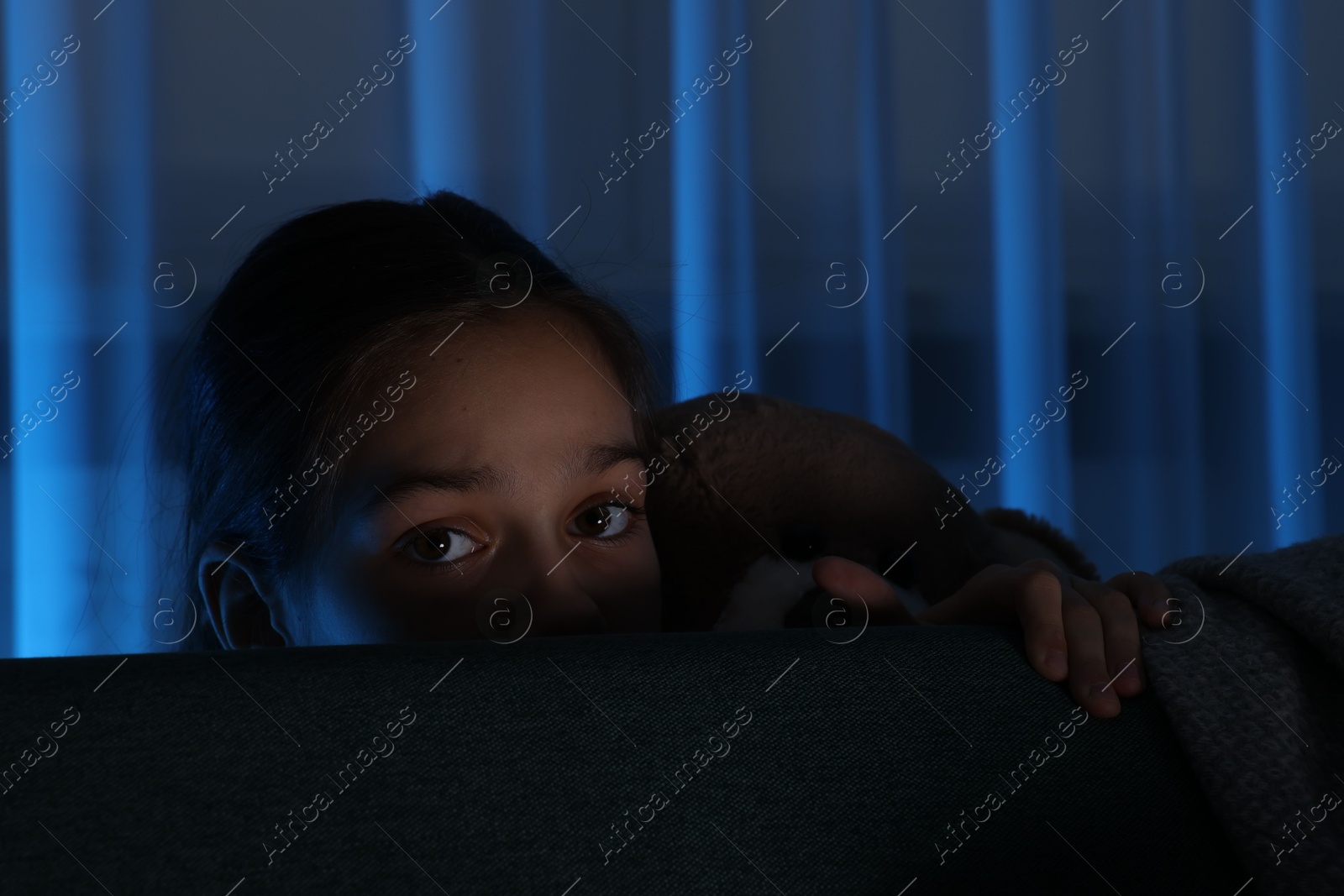 Photo of Scared girl with teddy bear hiding behind sofa at night