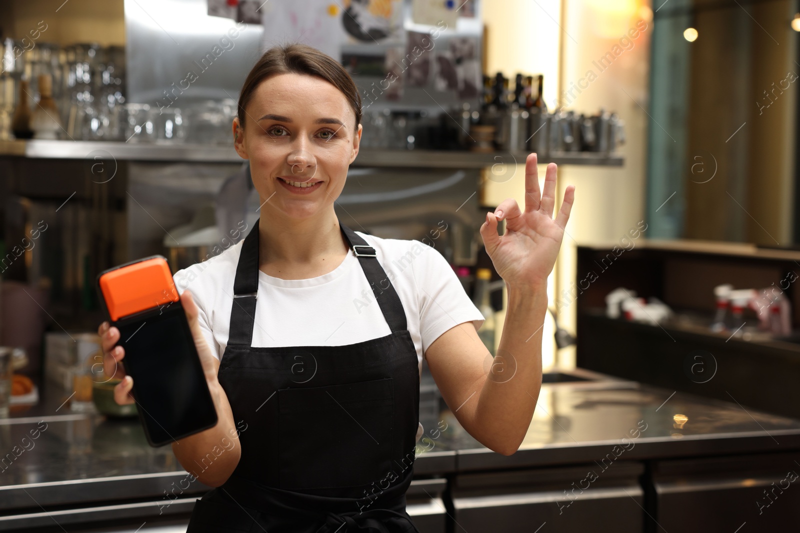 Photo of Smiling cafe worker with payment terminal showing OK gesture indoors