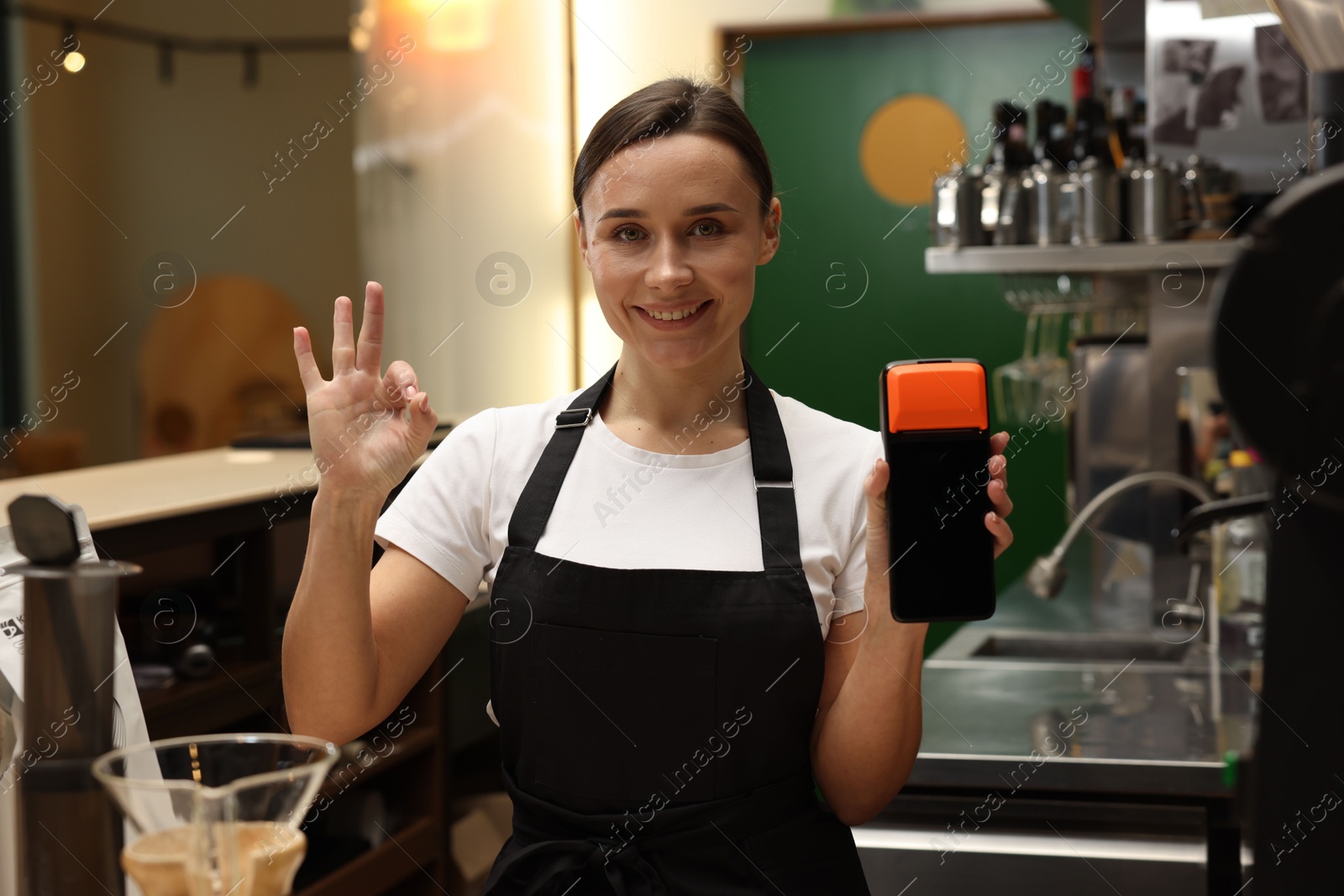 Photo of Smiling cafe worker with payment terminal showing OK gesture indoors