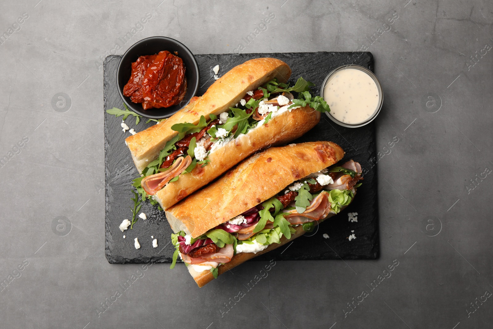 Photo of Halves of baguette sandwich with prosciutto and sun dried tomatoes served on grey table, top view