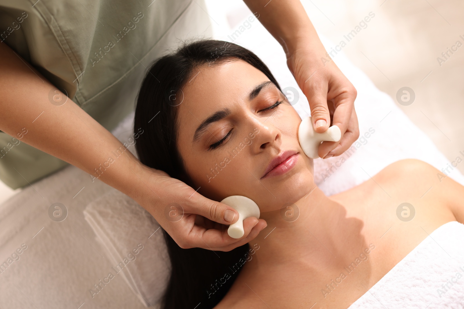 Photo of Young woman receiving facial massage with spa stones in salon