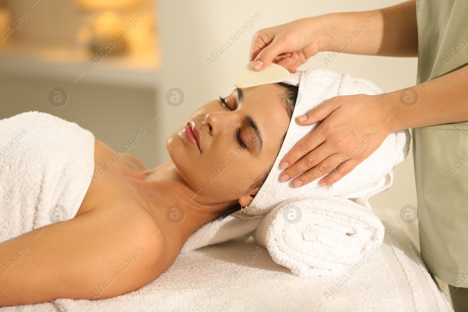 Photo of Young woman receiving facial massage with gua sha tool in spa salon