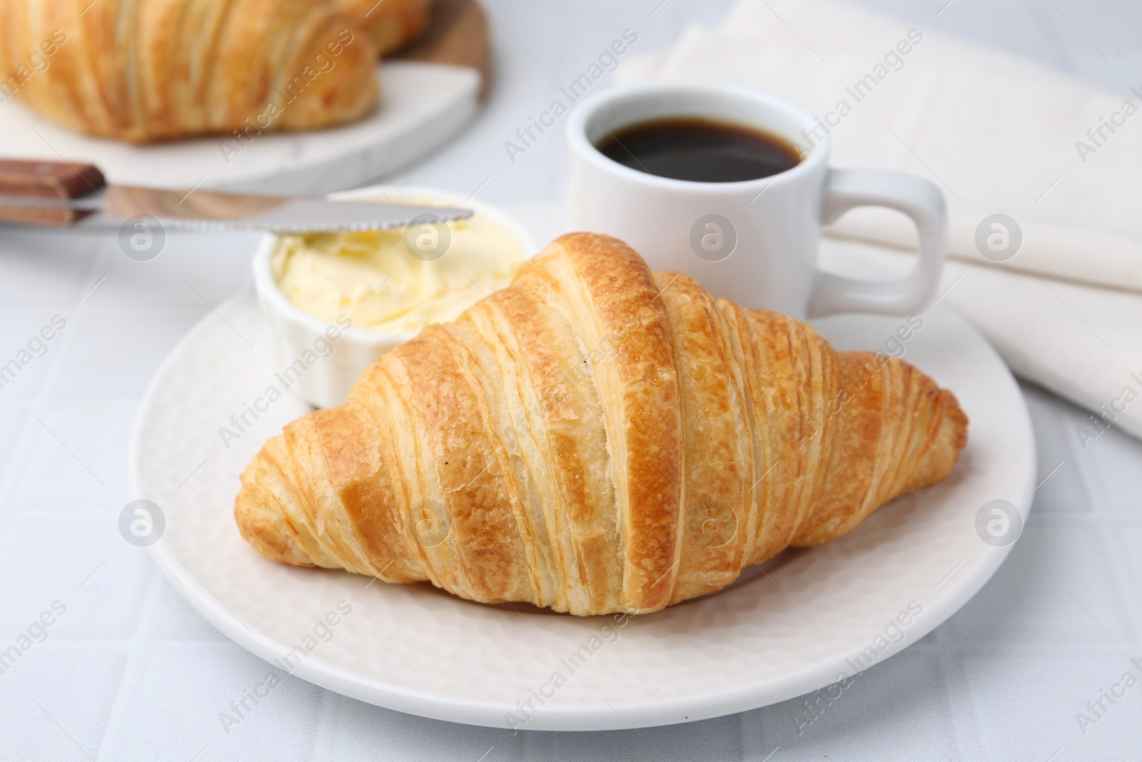 Photo of Tasty fresh croissant served with butter and cup of coffee on white tiled table, closeup