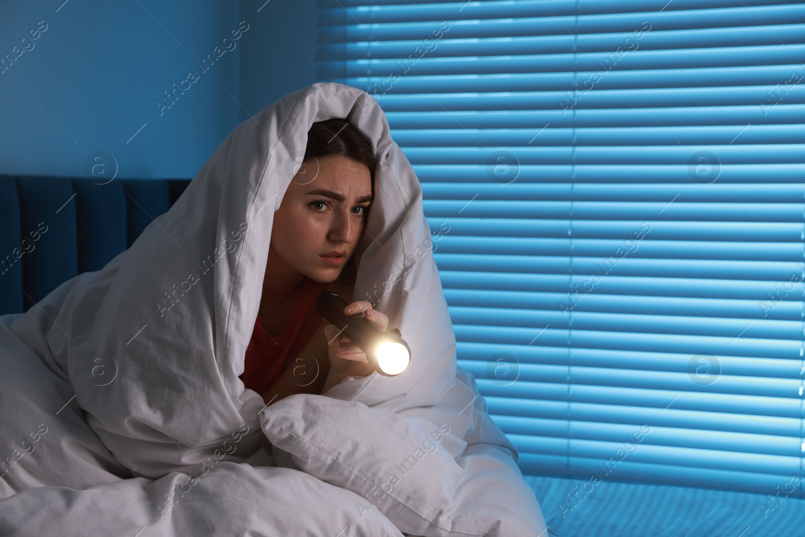 Photo of Fear of darkness. Scared young with blanket and flashlight on bed indoors at night, space for text