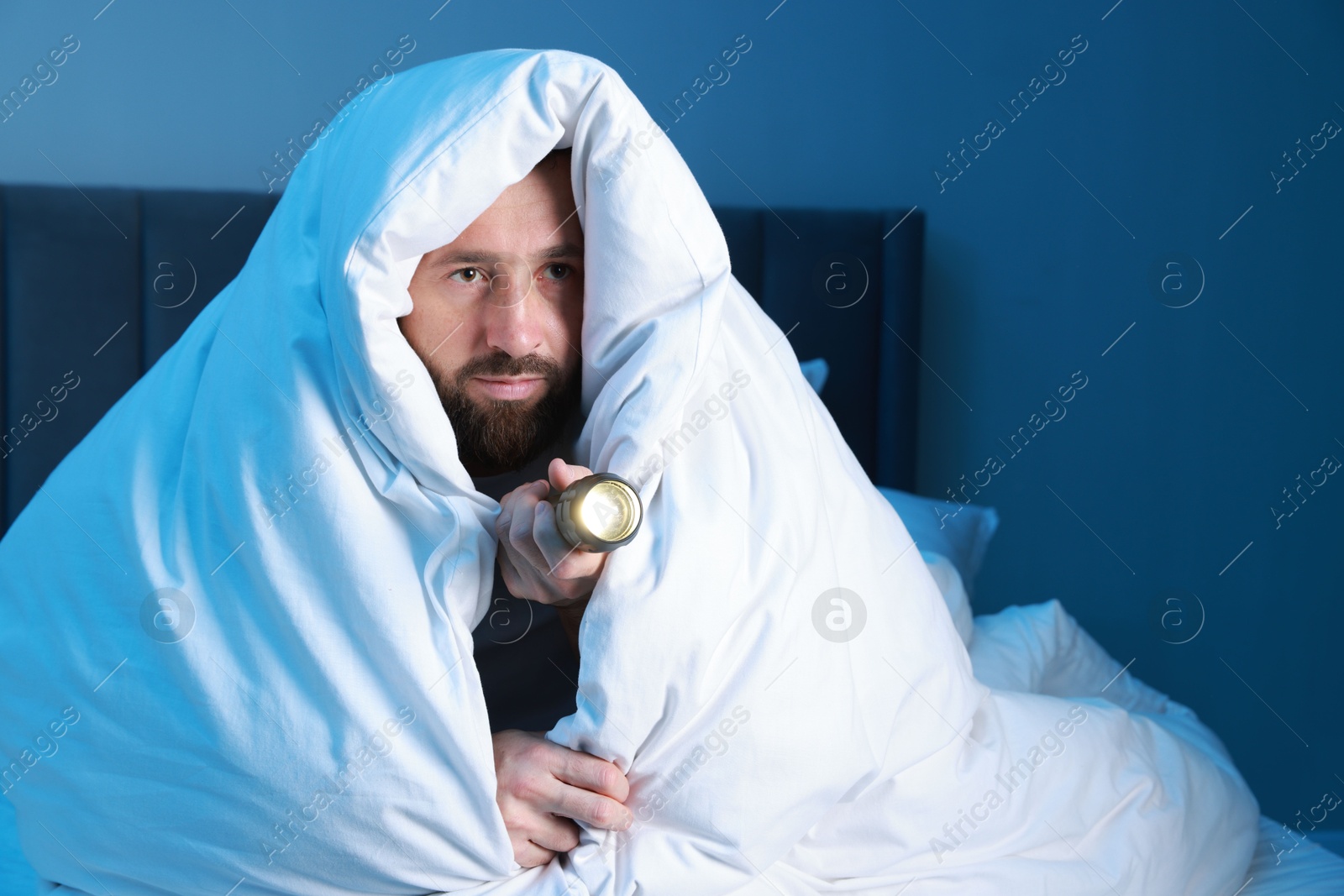 Photo of Fear of darkness. Scared man with blanket and flashlight on bed in room at night