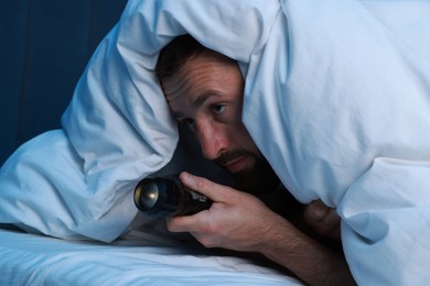 Photo of Fear of darkness. Scared man with blanket and flashlight on bed in room at night